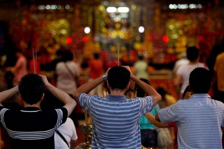 Worshippers praying at Singapore's Kwan Im Thong temple. The three men have their back to the camera and are facing the ornate altar. They are holding sticks of incense