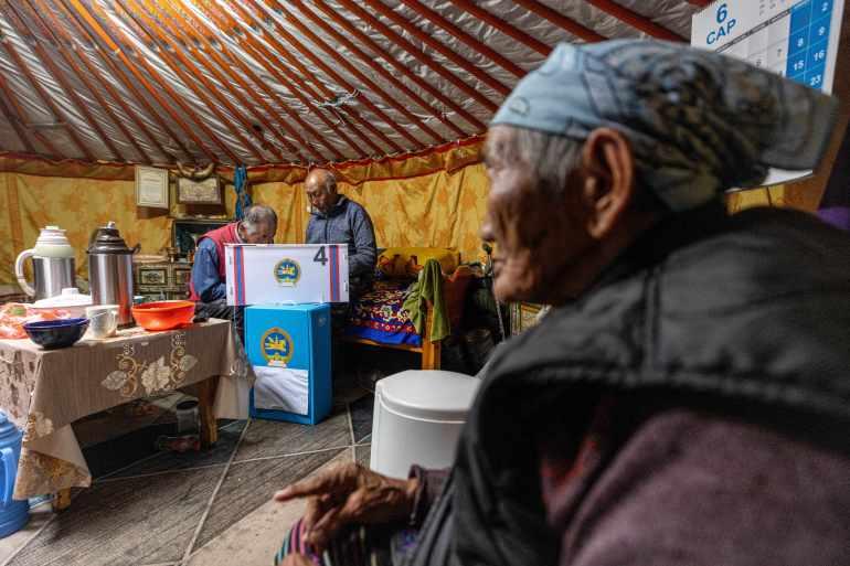 An older woman inside a traditional Mongolian gert. There is a mobile polling box set up. The woman has a scarf tied in her hair and is wearing a thick coat. Two elderly men are standing at the voting booth. There are flasks and bowls on a nearby table.